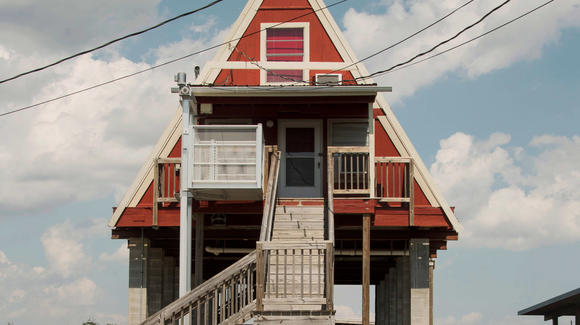Una casa de estilo A-frame en Luisiana, elevada sobre pilares de cemento, preparada para inundaciones. Con dos pisos, su fachada roja contrasta con el cielo parcialmente nublado. Al fondo, se ve el bayou.
